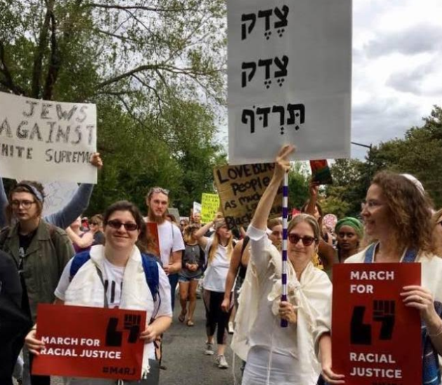 Photo credit: Susan Wasserkrug. The red signs was created by Lynna Schaefer. Carrying the red sign on the left: Rabbi Amber Powers. Carrying the Tsedek sign: Betsy Teutsch.  Carrying the red sign on the right, Lynna Schaefer
